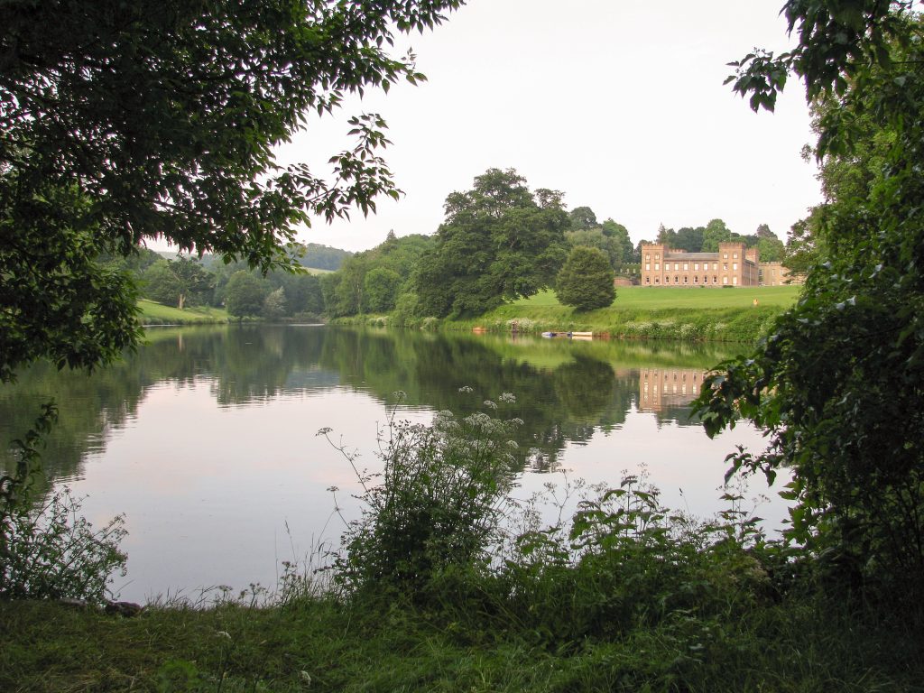 a beautiful stately home across a large lake, with vegetation growing around the egdes